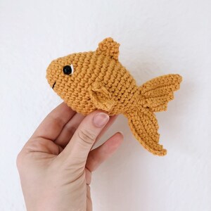 May include: A crocheted goldfish toy in a bright orange color. The toy has a black eye and a small, curled tail.