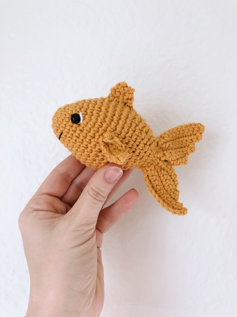 May include: A crocheted goldfish toy in a bright orange color. The toy has a black eye and a small, curled tail.