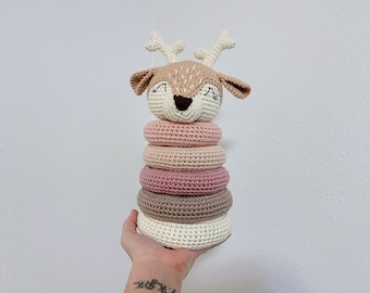 May include: A crocheted deer-shaped stacking toy with five rings in shades of pink, beige, and white. The deer has a smiling face with closed eyes.