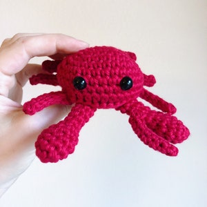 May include: A crocheted red crab with black eyes. The crab is small and has a cute, cartoon-like appearance.