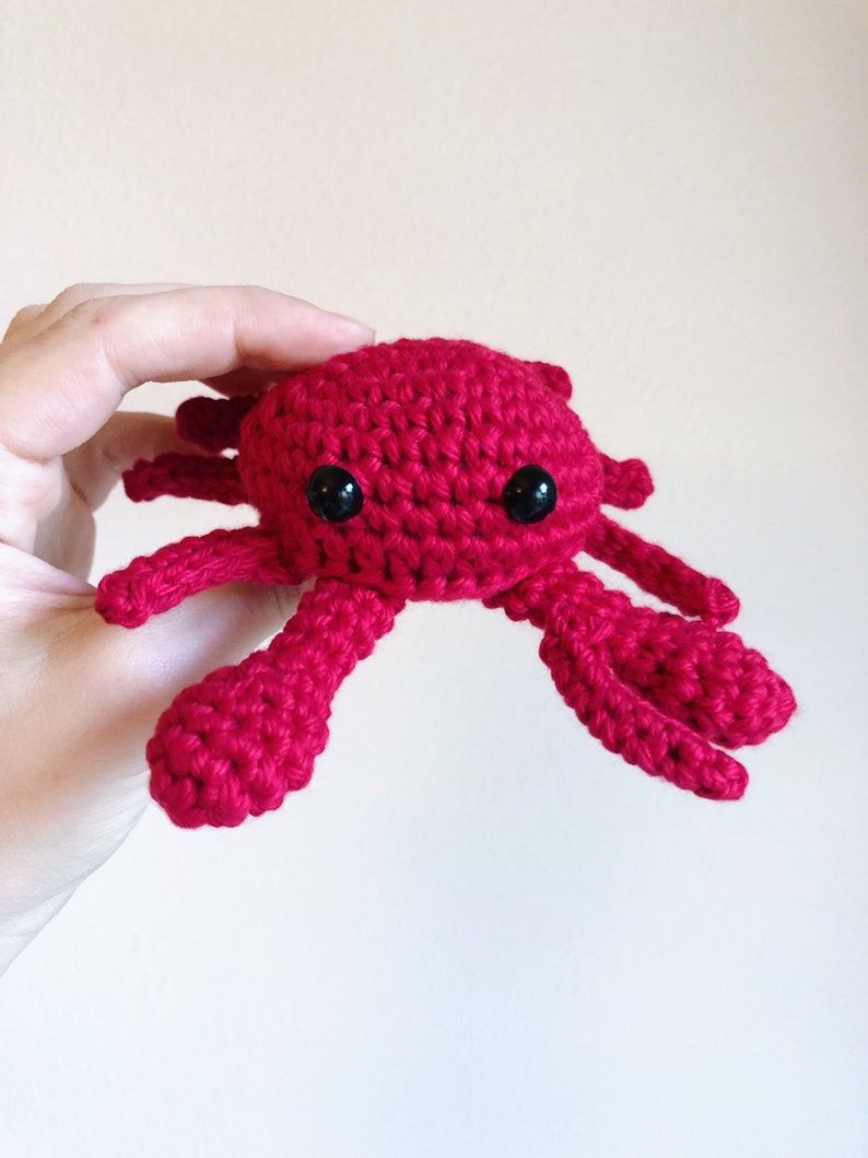 May include: A crocheted red crab with black eyes. The crab is small and has a cute, cartoon-like appearance.