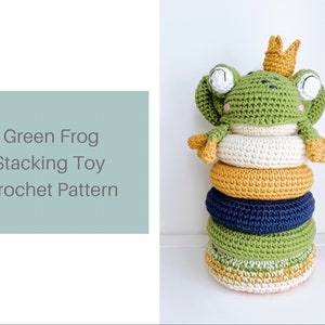 May include: Green crocheted frog stacking toy with a crown. The toy has five rings in different colours: white, yellow, navy blue, green, and a green ring with a yellow and white stripe.