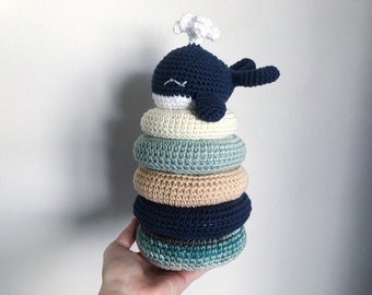 May include: A crocheted whale toy with a white spout sits atop a stack of five colorful crocheted rings. The rings are in shades of blue, green, and beige.