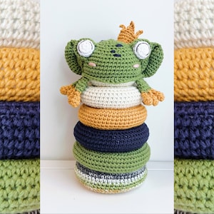 Könnte beinhalten: Ein grüner Häkel-Frosch mit einer goldenen Krone sitzt auf einem Stapel bunter Häkelringe. Die Ringe sind in den Farben Weiß, Gelb, Dunkelblau und Grün.