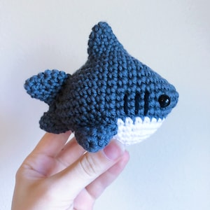 May include: A crocheted blue shark toy with white teeth and black eyes. The shark is being held by a hand.