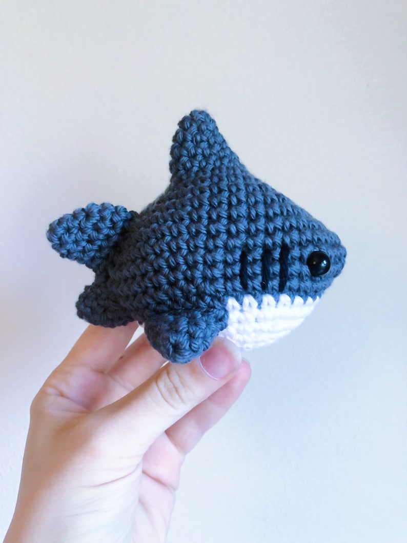 May include: A crocheted blue shark toy with white teeth and black eyes. The shark is being held by a hand.