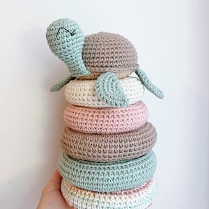 Puede incluir: Un juguete de apilar de tortuga de crochet con cinco anillos en tonos de verde, marrón, rosa y blanco. La tortuga tiene una cara sonriente y está sentada encima de los anillos.