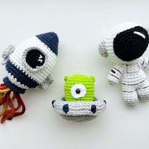 Puede incluir: Tres juguetes de peluche tejidos a crochet: un cohete blanco y azul con llamas naranjas, un extraterrestre verde en una nave espacial gris y un astronauta blanco con un casco negro.