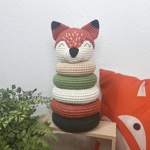 May include: A crocheted fox stacking toy with five rings in shades of green, beige, and red. The fox has black eyes and a black nose.