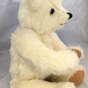 May include: A white plush teddy bear with black eyes and a brown nose. The bear is sitting with its legs crossed and its arms resting on its lap.