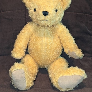 May include: A light brown teddy bear with a fluffy texture. The bear is sitting with its legs crossed and has a stitched nose and black eyes.