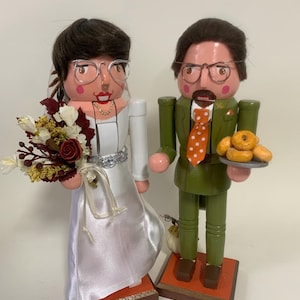 May include: Wooden nutcracker figurines of a bride and groom. The bride wears a white satin wedding dress and holds a bouquet. The groom wears a green suit and holds a plate of donuts. Both wear glasses and stand on wooden bases.