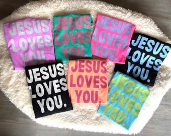 May include: Seven t-shirts with the text "Jesus Loves You" in different colors, including pink, turquoise, blue, black, and orange. The text is in a bold, blocky font.