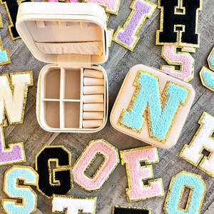 May include: A white jewelry box with a mirrored interior and multiple compartments. The box is surrounded by colorful felt letters with gold trim. The letters spell out the word "GOES"