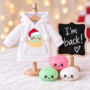 May include: A white long-sleeve hooded sweatshirt featuring a graphic of a green dumpling wearing a Santa hat. Beside it stands a small wooden chalkboard sign with the text I'm back! and a heart, along with three small round dumpling figurines.
