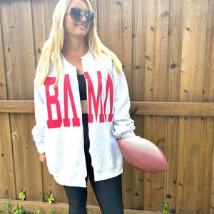 May include: A light gray zip-up sweatshirt with the word "BAMA" in large red block letters. The person is holding a brown football. The sweatshirt is paired with black leggings.