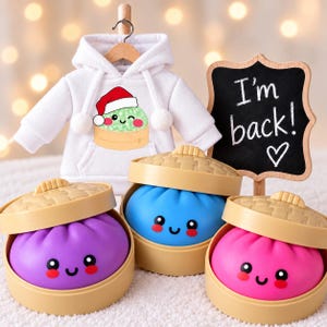 May include: A white hooded sweatshirt with a graphic of a dumpling wearing a Santa hat, accompanied by three round, colorful dumpling toys in bamboo steamers and a black chalkboard sign with the white text I'm back! and a heart symbol.