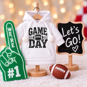 May include: A white hooded sweatshirt with the black text GAME DAY and a football graphic, a green foam finger with the text WE'RE #1, a brown football, and a black chalkboard sign with the white text Let's Go! and a heart symbol.