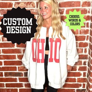 May include: White sweatshirt with the word "OHIO" in red block letters. The sweatshirt has a loose fit and is open in the front. The image also includes text that says "CUSTOM DESIGN" and "CHOOSE: WORDS & COLORS".
