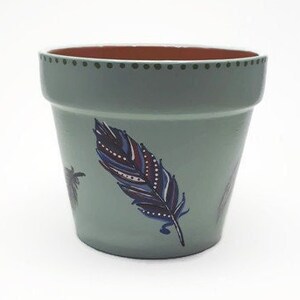 Peut inclure: Un pot en terre cuite vert clair avec un bord brun et un motif peint d'une plume bleue et violette avec des accents blancs.