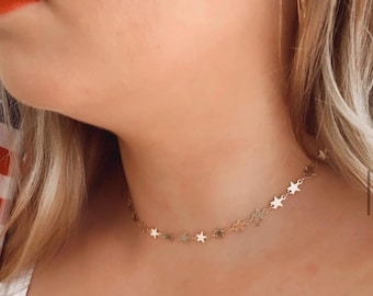 Może przedstawiać: Złoty naszyjnik typu choker z delikatnym łańcuszkiem ozdobionym serią małych, równomiernie rozmieszczonych zawieszek w kształcie gwiazdek. Naszyjnik przylega blisko szyi, prezentując prosty wzór ogniw z polerowanymi metalowymi akcentami gwiazd.
