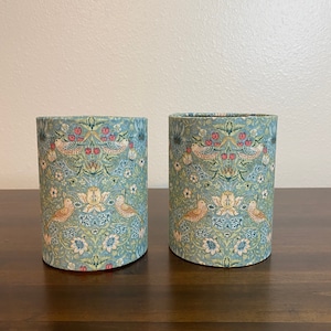 May include: Two cylindrical containers with a floral pattern featuring birds and leaves. The containers are made of a light blue fabric with a white background. The pattern is in shades of green, red, and yellow.