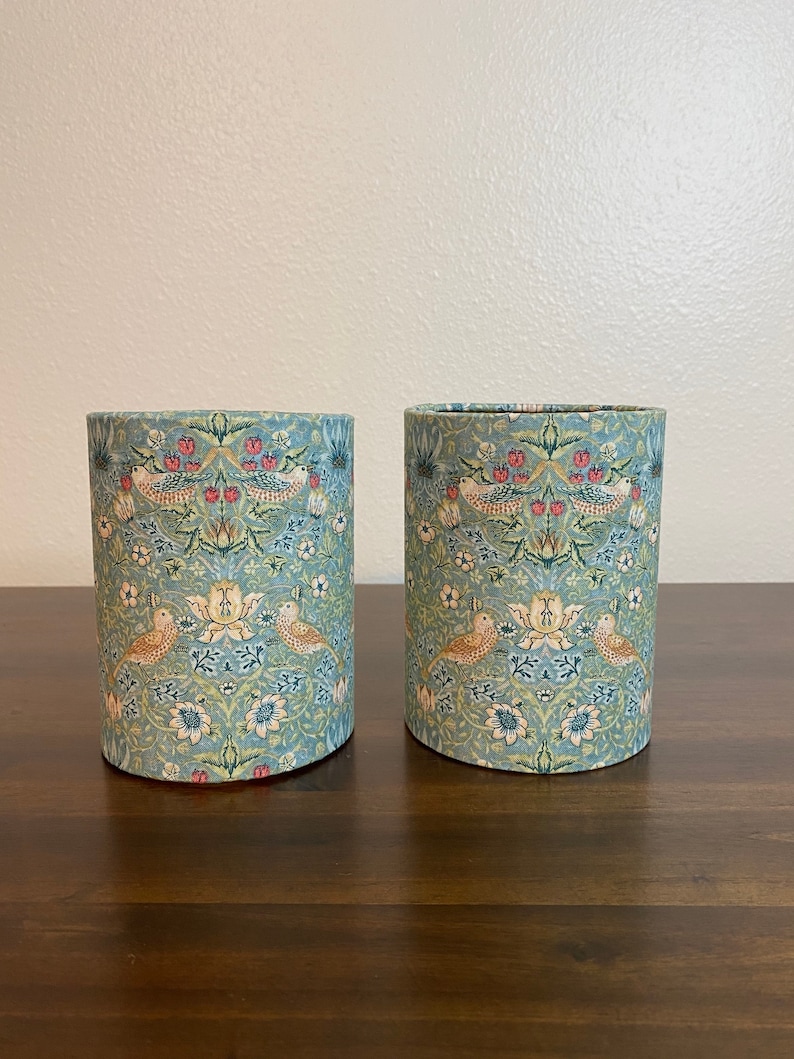 May include: Two cylindrical containers with a floral pattern featuring birds and leaves. The containers are made of a light blue fabric with a white background. The pattern is in shades of green, red, and yellow.