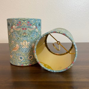 May include: Two lampshades with a floral pattern featuring birds and flowers in shades of blue, green, and pink. The lampshades are made of fabric and have a gold metal ring.