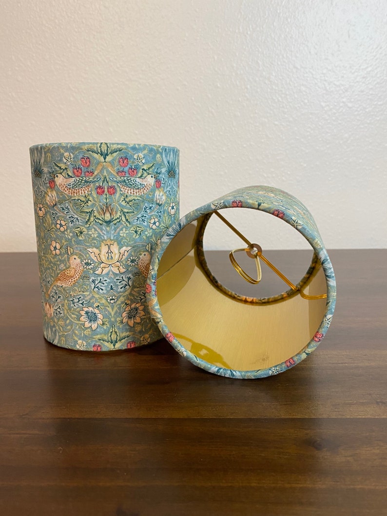 May include: Two lampshades with a floral pattern featuring birds and flowers in shades of blue, green, and pink. The lampshades are made of fabric and have a gold metal ring.