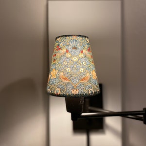 May include: A black metal wall-mounted lamp with a cone-shaped lampshade. The lampshade is covered in a floral pattern with blue, green, red, and yellow colors.