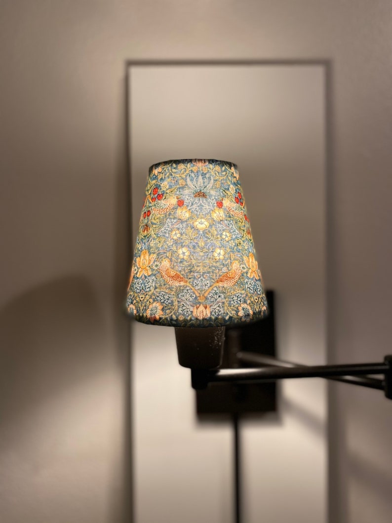 May include: A black metal wall-mounted lamp with a cone-shaped lampshade. The lampshade is covered in a floral pattern with blue, green, red, and yellow colors.