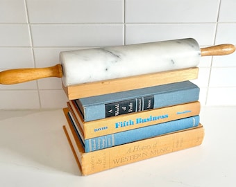 May include: A stack of books with a marble rolling pin on top. The books are various colors and have titles like "Fifth Business" and "A History of Western Music".
