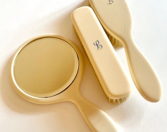 Può includere: Un set di tre articoli da toeletta color avorio: uno specchio da mano, una spazzola per capelli e una spazzola per vestiti. Ogni articolo ha un monogramma blu "LB" sul manico.