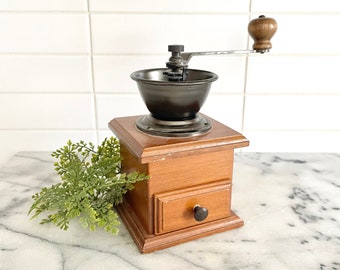 Puede incluir: Un molinillo de café vintage de madera con un acabado marrón oscuro. El molinillo tiene una manivela de metal y un cajón para guardar los granos de café. Está sentado en una encimera de mármol blanco.