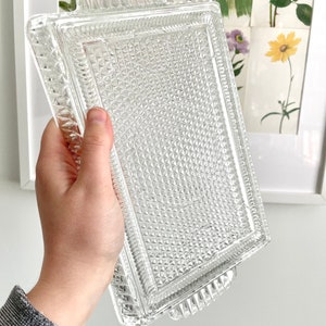 Puede incluir: Una bandeja rectangular de vidrio transparente con un patr&oacute;n de diamantes y asas decorativas. La bandeja se sostiene en una mano, mostrando su intrincado dise&ntilde;o. El fondo presenta estampados bot&aacute;nicos enmarcados.