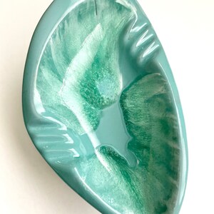 May include: A light green ceramic ashtray with a swirl design. The ashtray is shaped like a leaf and has a wide, shallow bowl.