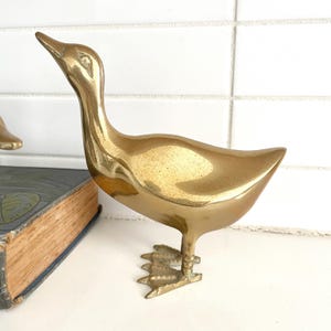 May include: A brass duck figurine with a long neck and detailed webbed feet. The duck is standing upright, with its head tilted upwards. The figurine has a shiny, golden finish and is likely a decorative item.
