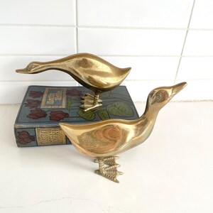 May include: Two brass duck figurines. One duck has its head down, the other has its head up. Both ducks are standing on brass feet. The ducks are on top of a blue book with red floral designs.