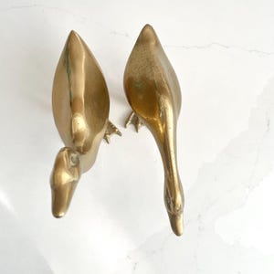May include: Two vintage brass swan figurines. The swans are depicted with their necks curved downward, and their wings are folded. The brass has a warm, golden hue, and the details of the feathers and beaks are visible.
