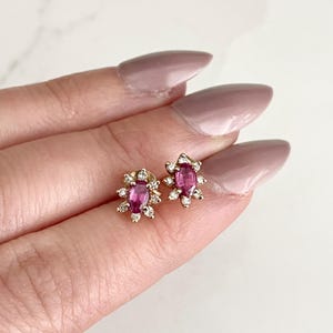 Könnte beinhalten: Ein Paar goldene Ohrringe mit ovalen Rubin-Edelsteinen, die von einem Heiligenschein aus kleineren Diamanten umgeben sind.