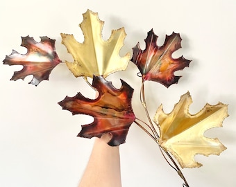 May include: A wall sculpture featuring five metal leaves in shades of gold, copper, and brown. The leaves are arranged in a cluster and have a textured, hammered finish.