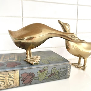 May include: Two brass duck figurines. One duck is bending its neck down, and the other is standing upright. The ducks are on a vintage book with a floral design. The ducks are a shiny gold color.