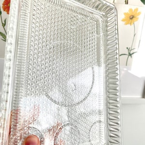Puede incluir: Bandeja de servir rectangular de vidrio transparente con un patr&oacute;n de diamantes y tres hendiduras circulares. La bandeja tiene una superficie texturizada y un borde decorativo. La bandeja es sostenida por una persona.