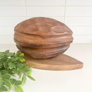 Puede incluir: Un cuenco de madera tallada con tapa, con un diseño texturizado y geométrico. El cuenco es de color marrón cálido y se apoya sobre una base de madera en forma de hoja. Decorativo y funcional.