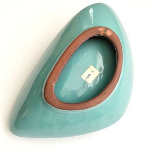 May include: A teal blue ceramic bowl with a brown rim. The bowl is shaped like a teardrop and has a small label on the bottom that says "Made in".