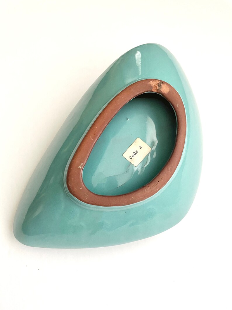 May include: A teal blue ceramic bowl with a brown rim. The bowl is shaped like a teardrop and has a small label on the bottom that says "Made in".
