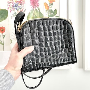 May include: Black handbag with a crocodile-effect texture. The bag features a gold-coloured clasp and a long, thin strap. The purse is held by a person wearing a grey sweater.