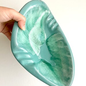 May include: A light blue ceramic ashtray with a green swirl design. The ashtray is shaped like a leaf and has a wide, shallow bowl.