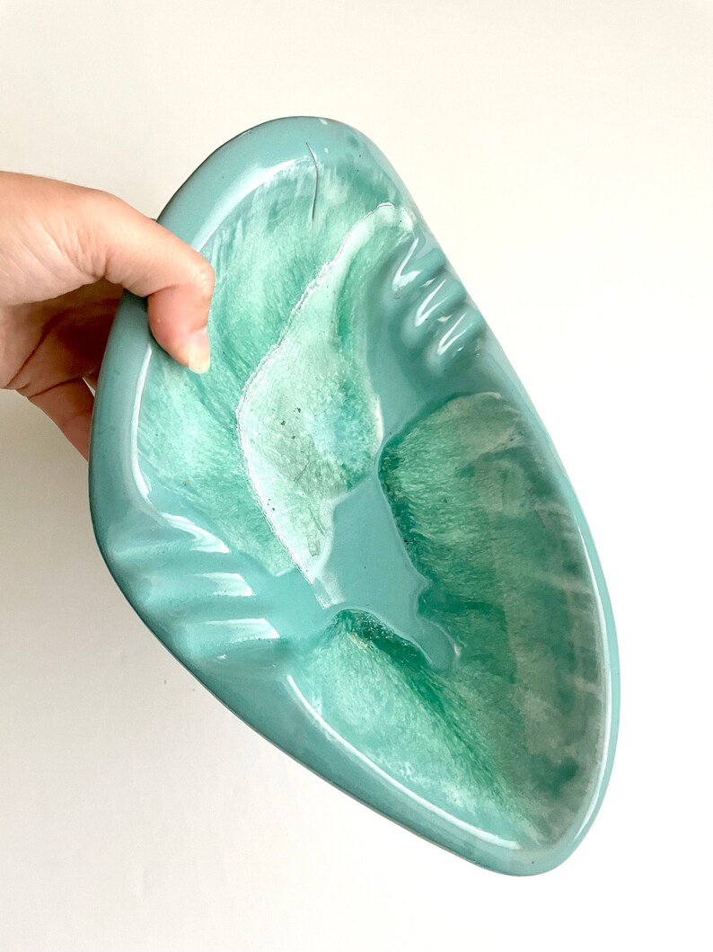 May include: A light blue ceramic ashtray with a green swirl design. The ashtray is shaped like a leaf and has a wide, shallow bowl.