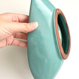 May include: A light blue ceramic planter with a rounded, oval shape and a brown rim. The planter is designed to be hung on a wall.